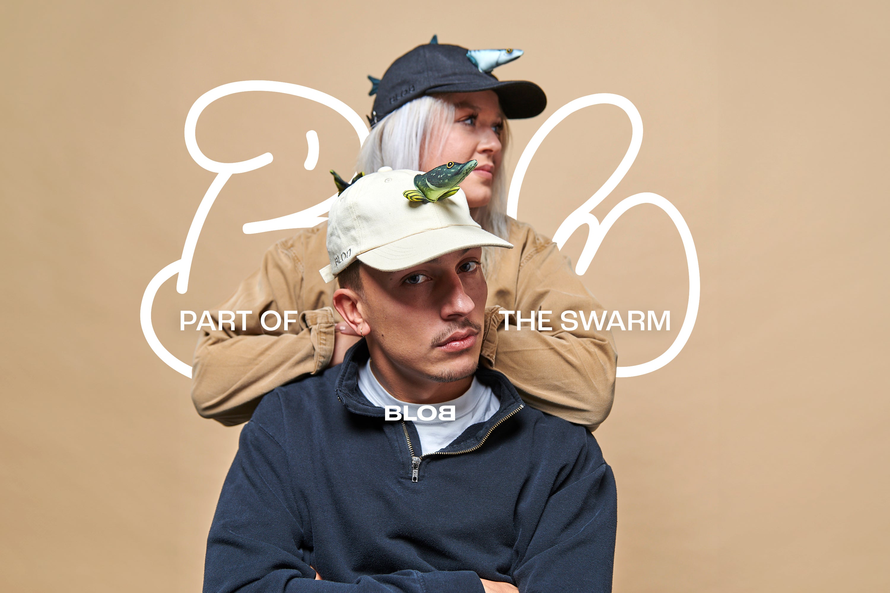 Zwei Personen tragen stylische Kappen mit 3D-Fischdesign. Eine Person steht im Hintergrund mit dunkler Fischmütze, die andere sitzt vorne mit heller Fischmütze. Im Hintergrund ist ein beiger Studiohintergrund mit großem „BLOB“-Branding und dem Text „PART OF THE SWARM“.